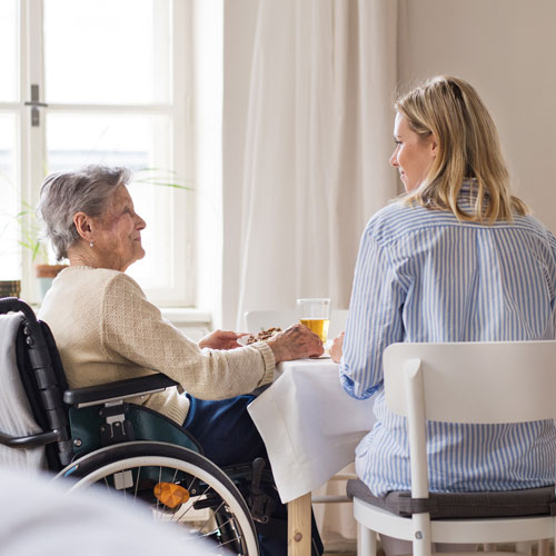 une femme discutant avec une personne âgée en fauteuil roulant autour d'une table, en train de prendre son repas