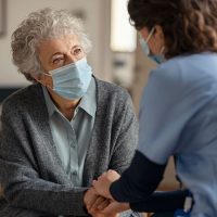 Aide soignante qui tient la main à une personne agée qui porte un masque